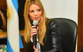 Imagen de Beatriz Domingorena con la bandera Argentina de fondo. Ella está sentada tras un escritorio con un micrófono en la mano